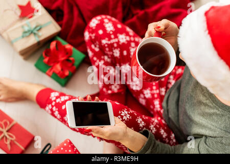Young woman wearing santa hat and xmas pajamas sitting on the floor amongst wrapped christmas presents, drinking hot punch and shopping online on her  Stock Photo