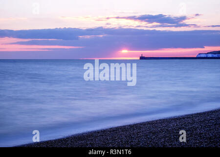 The sea is calm with movement, in the distance there is a harbour wall with a lighthouse on the end of it behind are some cliffs, the sky has scattere Stock Photo