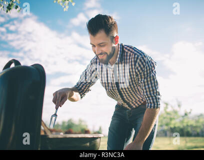 Handsome man preparing barbecue for friends. man cooking meat on barbecue - Chef putting some sausages and pepperoni on grill in park outdoor - Concep Stock Photo