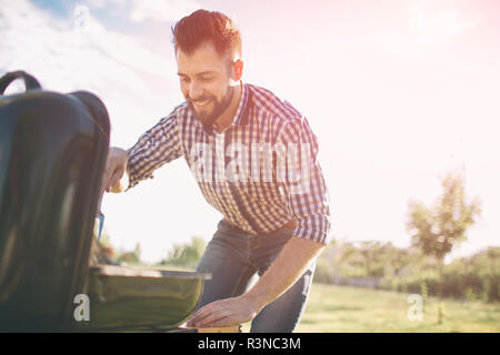 Handsome man preparing barbecue for friends. man cooking meat on barbecue - Chef putting some sausages and pepperoni on grill in park outdoor - Concep Stock Photo