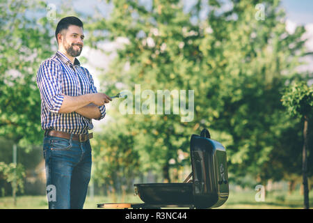 Handsome man preparing barbecue for friends. man cooking meat on barbecue - Chef putting some sausages and pepperoni on grill in park outdoor - Concep Stock Photo