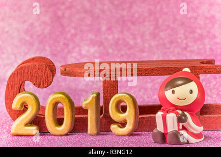 Pink glitter background for New Year's Cards with cute figurine of child in Christmas clothes with a present box in hands above a wooden sled and hand Stock Photo