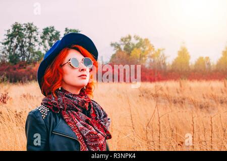 Beautiful red-haired woman in blue hat, dark round glasses and leather jacket walking in the park in the weather in the fall.  Copy space for text. Stock Photo