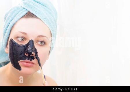 Woman in bathroom to put on cosmetic black mask on white background, front view. Sunny day. Put yourself in order. Stock Photo