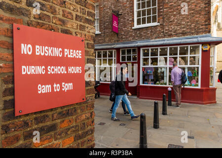 People pass red sign attached to exterior brick wall, polite request saying 'no buskers please during school hours'  - York, Yorkshire, England, UK. Stock Photo