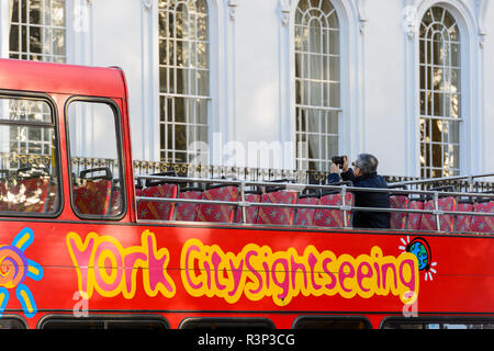 A solitary male tourist passenger photographs the De Grey Rooms from the top deck of red City Sightseeing bus - York, North Yorkshire, England, UK. Stock Photo