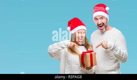 Middle age hispanic couple wearing christmas hat and holding gift over isolated background very happy pointing with hand and finger Stock Photo