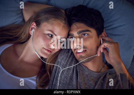 Young couple lying on cushions, sharing earphones, listening music