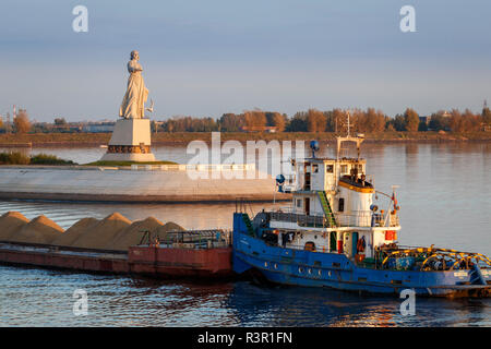 The statue Mother Volga, with hand outstreched in welcome, in the Rybinsk reservoir on the Volga River. A barge passes with mined raw materials. Stock Photo