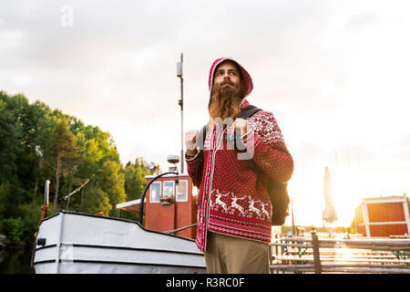 Finland, Kajaani, Cheerful young man. wearing houded jacket, standing on jetty, carrrying rucksack Stock Photo