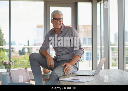 Portrait of smiling mature man with laptop sitting on table at home Stock Photo