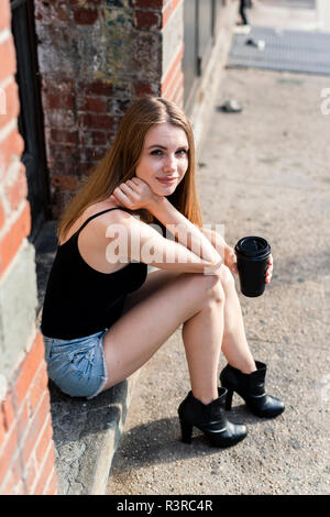 Young woman sitting in the street on a doorstep, holding a cup of coffee Stock Photo