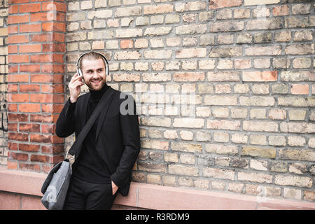 Smiling fashionable young man with headphones leaning against brick wall Stock Photo