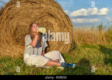 A girl with long hair sits on the ground in a field near a haystack and drinks milk from a large bottle Stock Photo