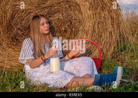 A girl with long hair sits on the ground in a field near a haystack and drinks milk from a large bottle Stock Photo
