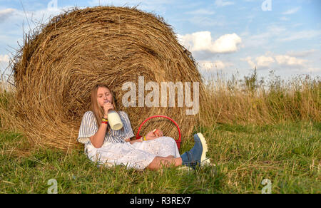 A girl with long hair sits on the ground in a field near a haystack and drinks milk from a large bottle Stock Photo