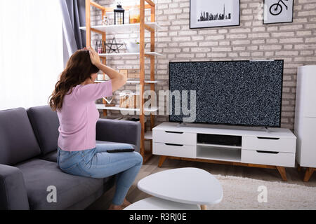 Frustrated Young Woman Sitting On Sofa Looking At Television With No Signal Stock Photo