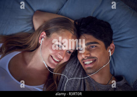 Young couple lying on cushions, sharing earphones, listening music Stock Photo