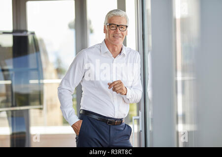 Businessman in office leaning against window, smiling Stock Photo