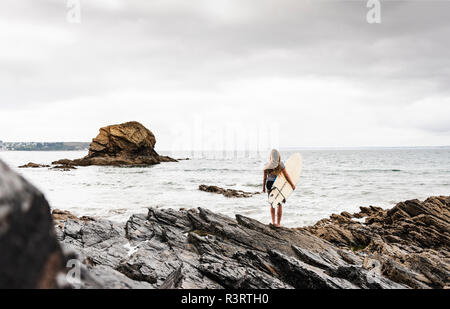 Young woman carrying surfboard on a rocky beach at the sea Stock Photo