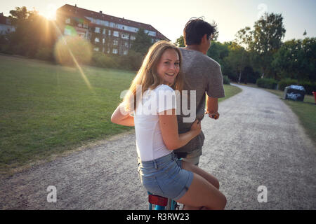 Young couple riding bicycle in park, woman sitting on rack Stock Photo