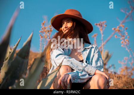 Smiling young woman wearing a hat sitting at an agave in the countyside Stock Photo