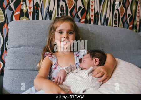Smiling girl sitting on couch holding newborn baby brother Stock Photo