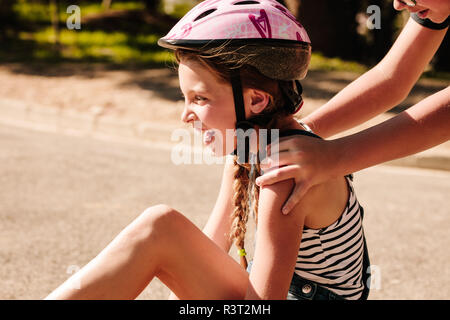 Girl sitting on street wearing a protective helmet with a boy pushing her from behind. Cropped shot of kids playing on street. Stock Photo