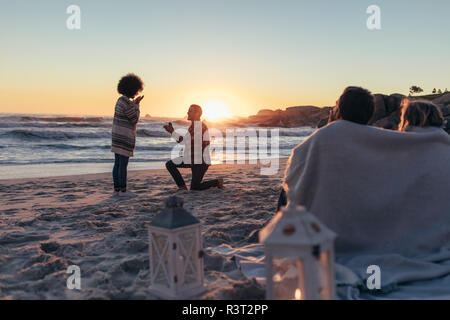 Man proposing woman her loved at sea shore. Marriage proposal at sunset beach with friends sitting in front wrapped in a blanket. Stock Photo