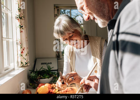 Cheerful old woman cutting vegetables while her husband is cooking food. Elderly couple having fun cooking food together at home. Stock Photo