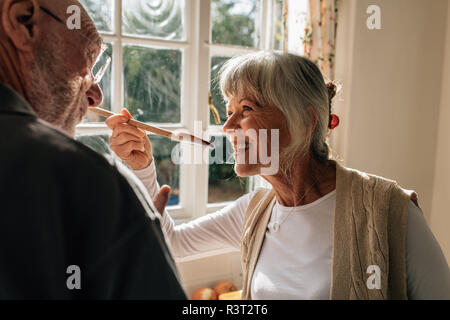 Senior woman tasting food using a wooden spoon in kitchen. Smiling woman standing in kitchen with her husband checking the taste of food. Stock Photo