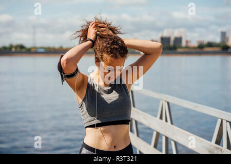Young athletic woman wearing earbuds at the riverside making her hair Stock Photo