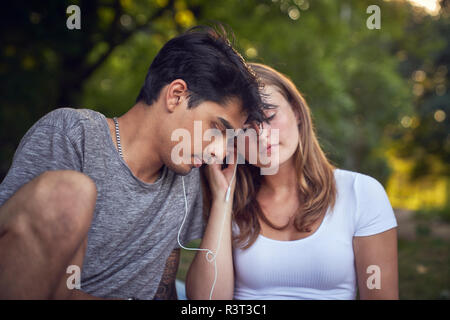 Young couple sitting in park, sharing earphones, listening music Stock Photo