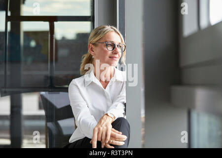 Serene businesswoman sitting on ground, looking out of window Stock Photo