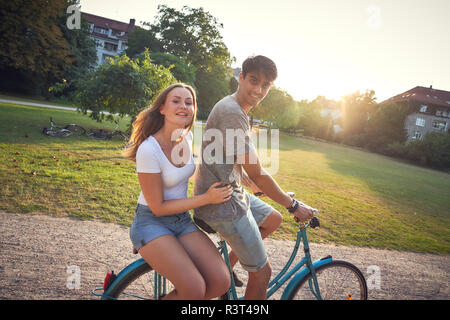 Young couple riding bicycle in park, woman sitting on rack Stock Photo
