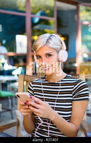 Portrait of smiling young woman with smartphone listening music with headphones at pavement cafe Stock Photo