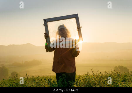 Italy, Tuscany, Borgo San Lorenzo, senior man holding window frame in field at sunrise above rural landscape Stock Photo