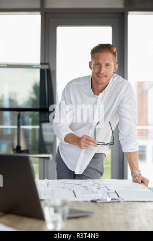 Portrait of smiling young man working on blueprint on desk in office Stock Photo