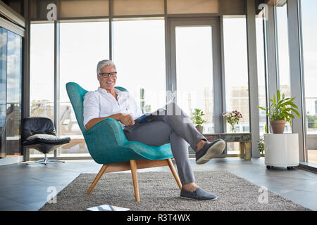 Portrait of smiling mature man with a brochure relaxing in armchair at the window at home Stock Photo