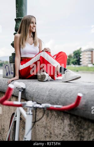 Smiling teenage girl sitting on a wall at the riverside next to bicycle Stock Photo