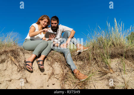 Young couple sitting on a dune in summer, using digital tablet Stock Photo