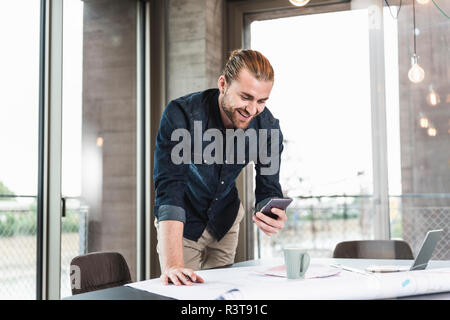 Smiling young businessman at desk in office with plan and cell phone Stock Photo