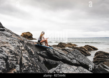 Young woman with surfboard sitting on the beach, using smartphone Stock Photo