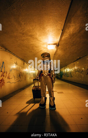 Spaceman in the city at night standing in underpass with rolling suitcase Stock Photo