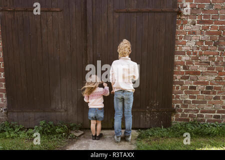 Back view of brother and his little sister standing in front of wooden door Stock Photo