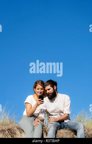 Young couple sitting on a dune in summer, using smartphone Stock Photo