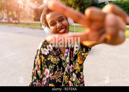 Happy fashionable young woman with headphones outdoors at sunset making victory gesture Stock Photo