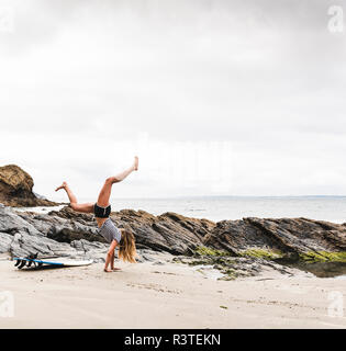 Young woman with surfboard doing handstand on the beach Stock Photo