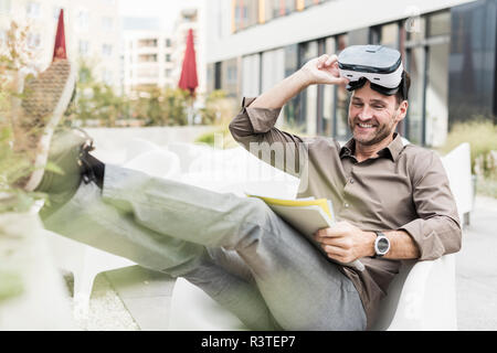 Smiling man with Virtual Reality Glasses and documents sitting on terrace Stock Photo