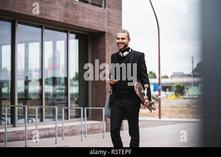 Smiling fashionable young man holding cell phone and skateboard passing office building Stock Photo
